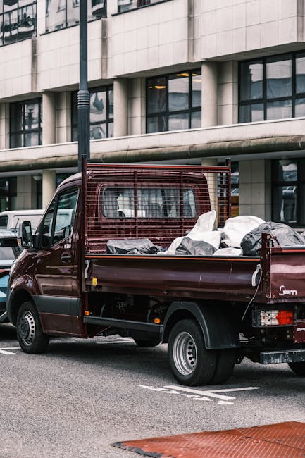 A small burgundy pickup truck parked on an urban street in front of a modern building with large rectangular windows and a concrete facade. The truck's open flatbed is filled with various waste materi