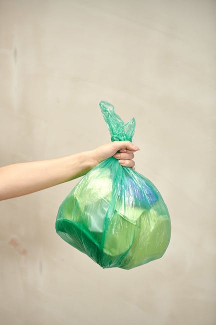 A person's hand is visible holding a green plastic rubbish bag tied at the top with a twist tie. The bag appears to be filled with waste and has a slightly crumpled texture with varying shades of gree