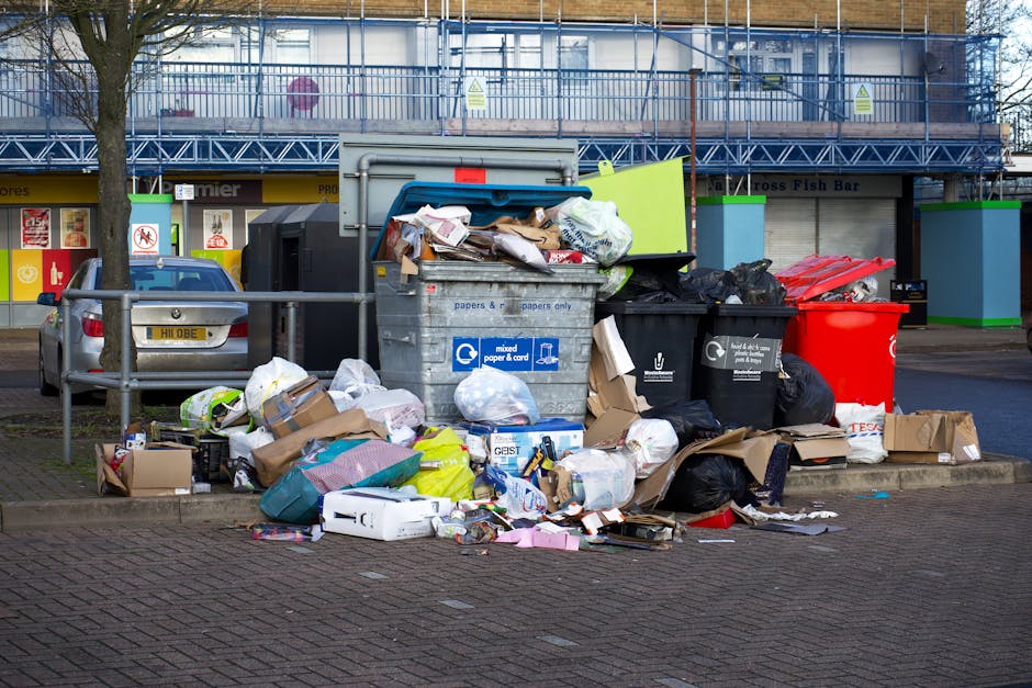 A person's hand is visible holding a green plastic rubbish bag tied at the top with a twist tie. The bag appears to be filled with waste and has a slightly crumpled texture with varying shades of gree